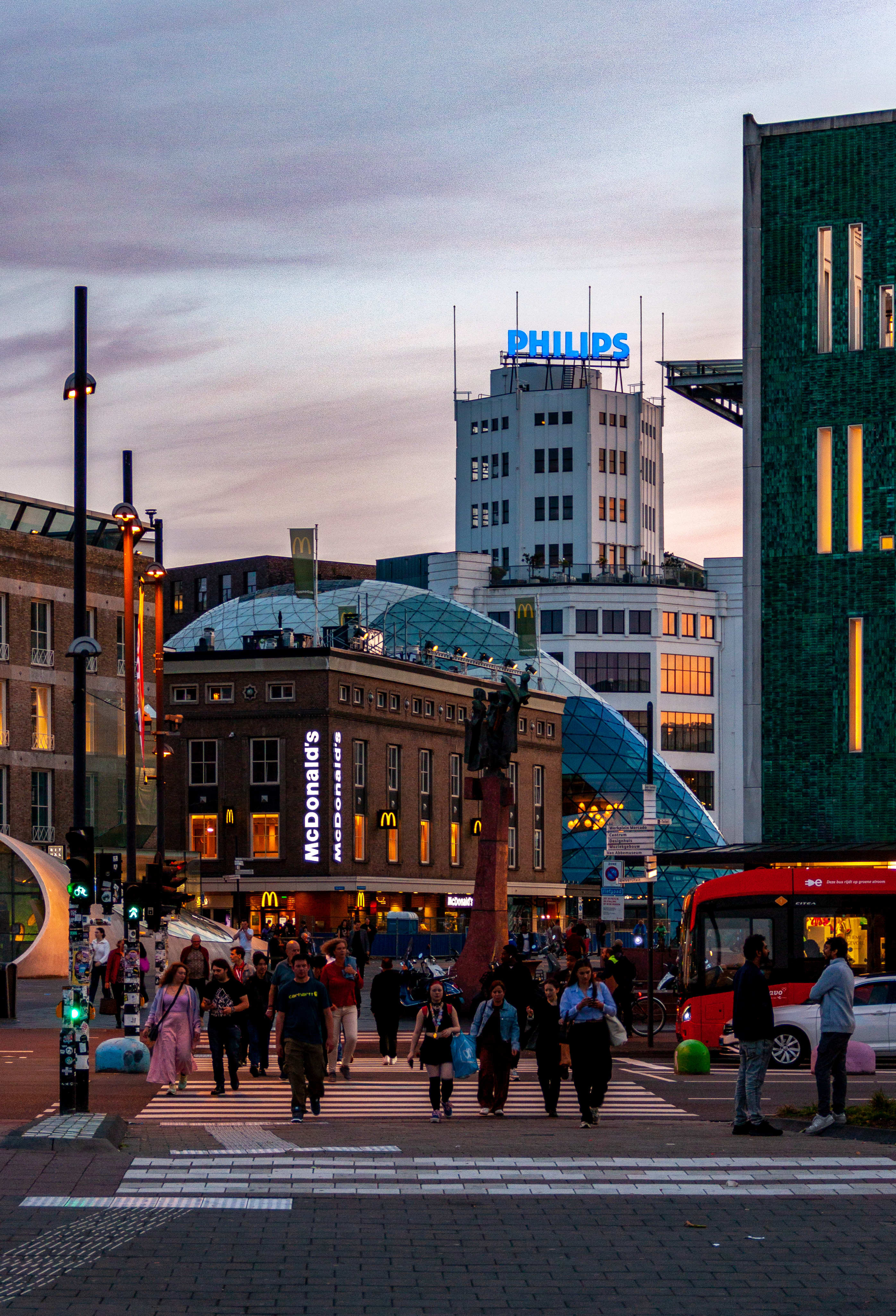 A shot of the 18-Septemberplein in Eindhoven during sunset. In the foreground there is people crossing the street at a crosswalk. In the background there is a McDonald's and an old factory with an illuminated Philips sign on the roof.