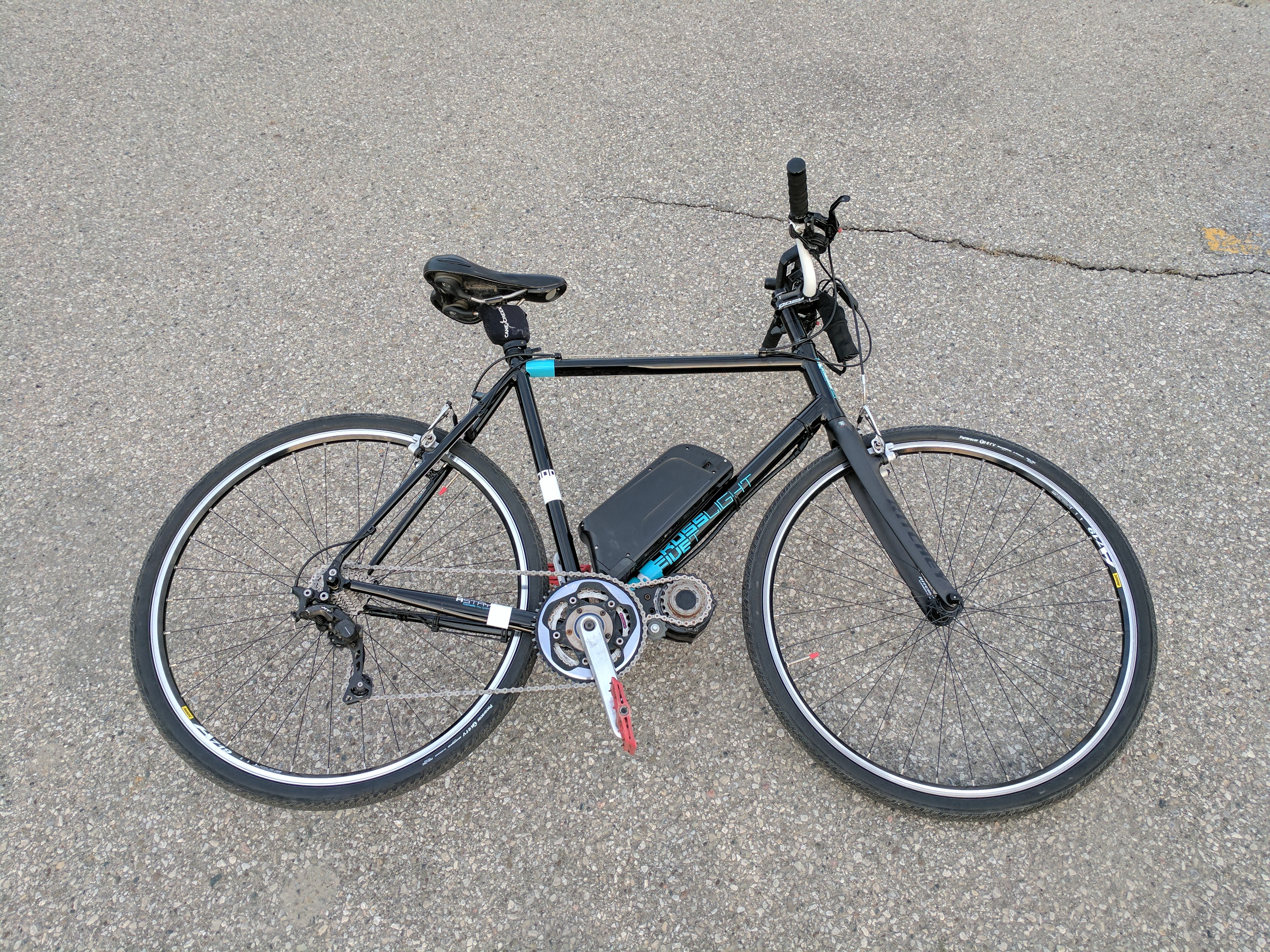  Black electric bicycle on gray asphalt pavement, featuring a battery pack mounted on the frame, front and rear wheels with silver rims, black handlebars with brake levers, a black saddle, and visible gear mechanism.