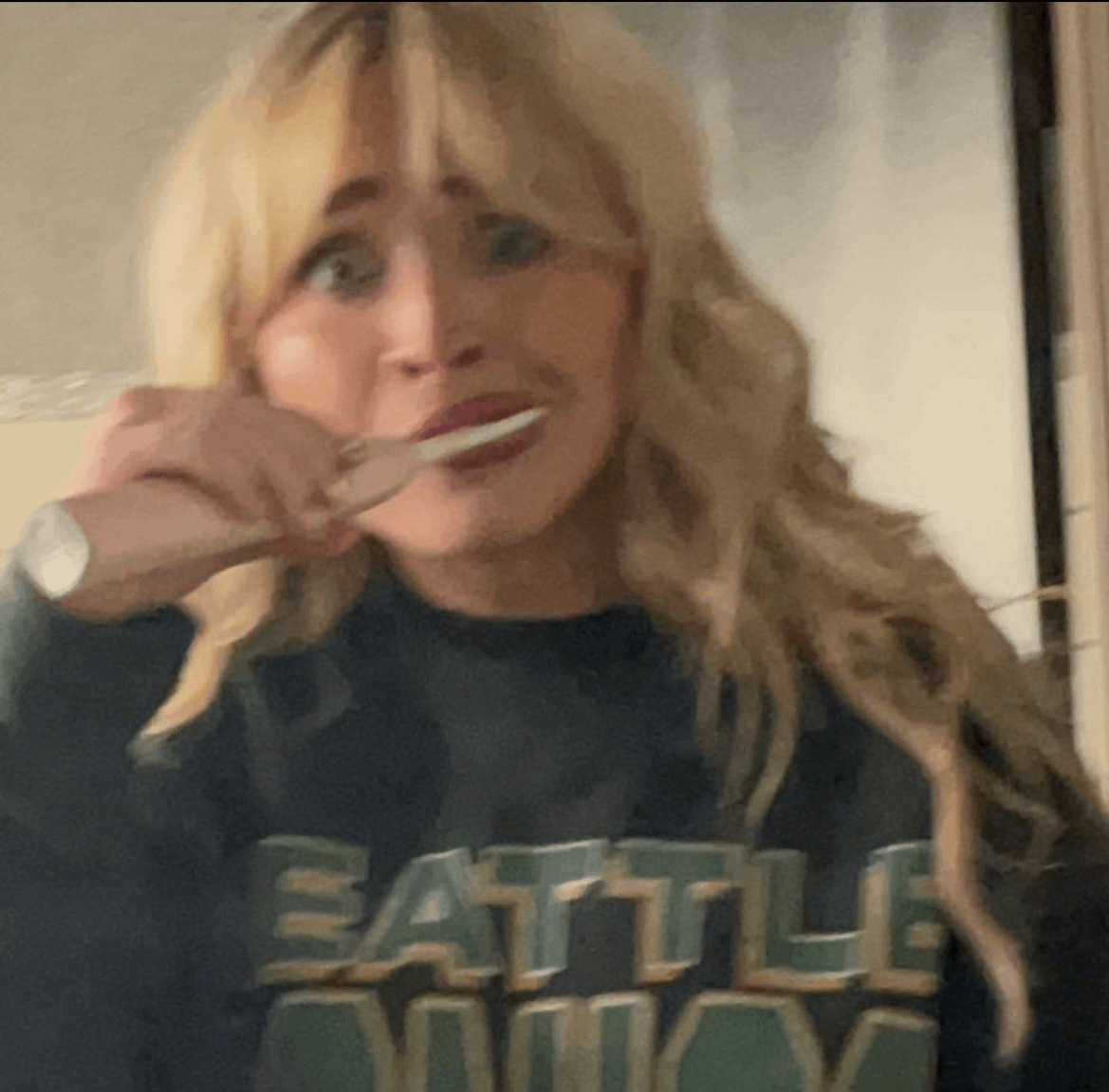 A grainy, slightly blurry selfie of a woman with blonde, wavy hair and bangs. She is wearing a dark navy or black sweatshirt with "SEATTLE" written across the chest in large, blocky green letters. She has an wide-eyed, humorous expression while holding a pink electric toothbrush to her mouth as if brushing her teeth.
