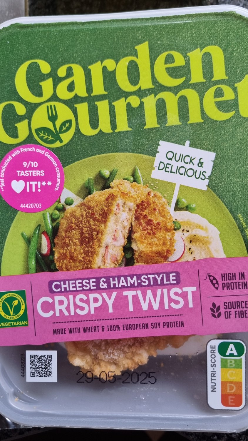 Garden Gourmet Cheese & Ham-Style Crispy Twist package showing a vegetarian frozen food product in a clear plastic tray. The green and yellow branded packaging displays a golden-brown breaded cylindrical food item cut in half to reveal a cheese and ham-style filling inside. The package includes a pink "9/10 TASTERS LOVE IT" badge, "QUICK & DELICIOUS" label, and indicates the product is high in protein, a source of fiber, and made with wheat and 100% European soy protein. A Nutri-Score A rating and vegetarian symbol are visible.
